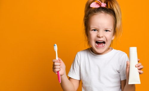 Cheerful girl with a toothbrush and toothpaste looks into the camera in yellow background
