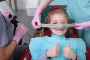 A little girl is comfortable to treat her teeth under superficial sedation. The girl smiles and holds two thumbs up. Milk teeth treatment. Treatment of children's teeth with nitrous oxide.